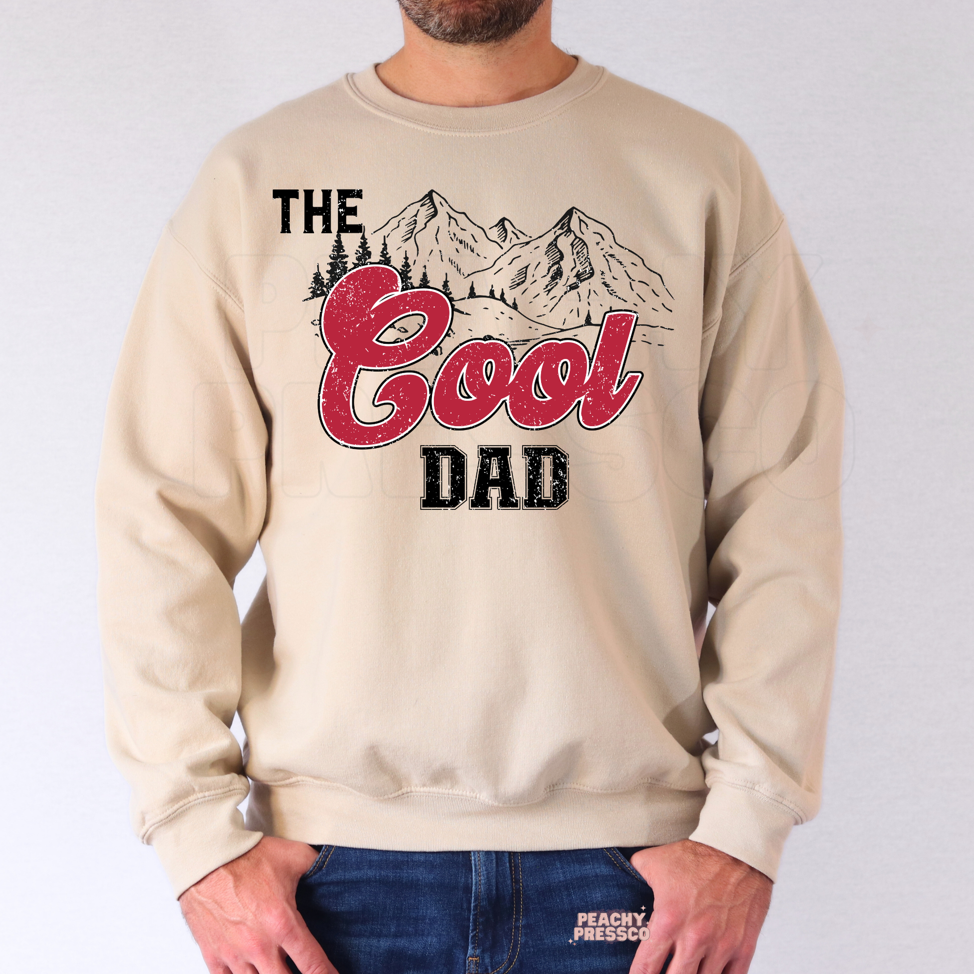 Man wearing beige crewneck sweatshirt featuring distressed “The Good Dad” graphic with mountain scenery design.