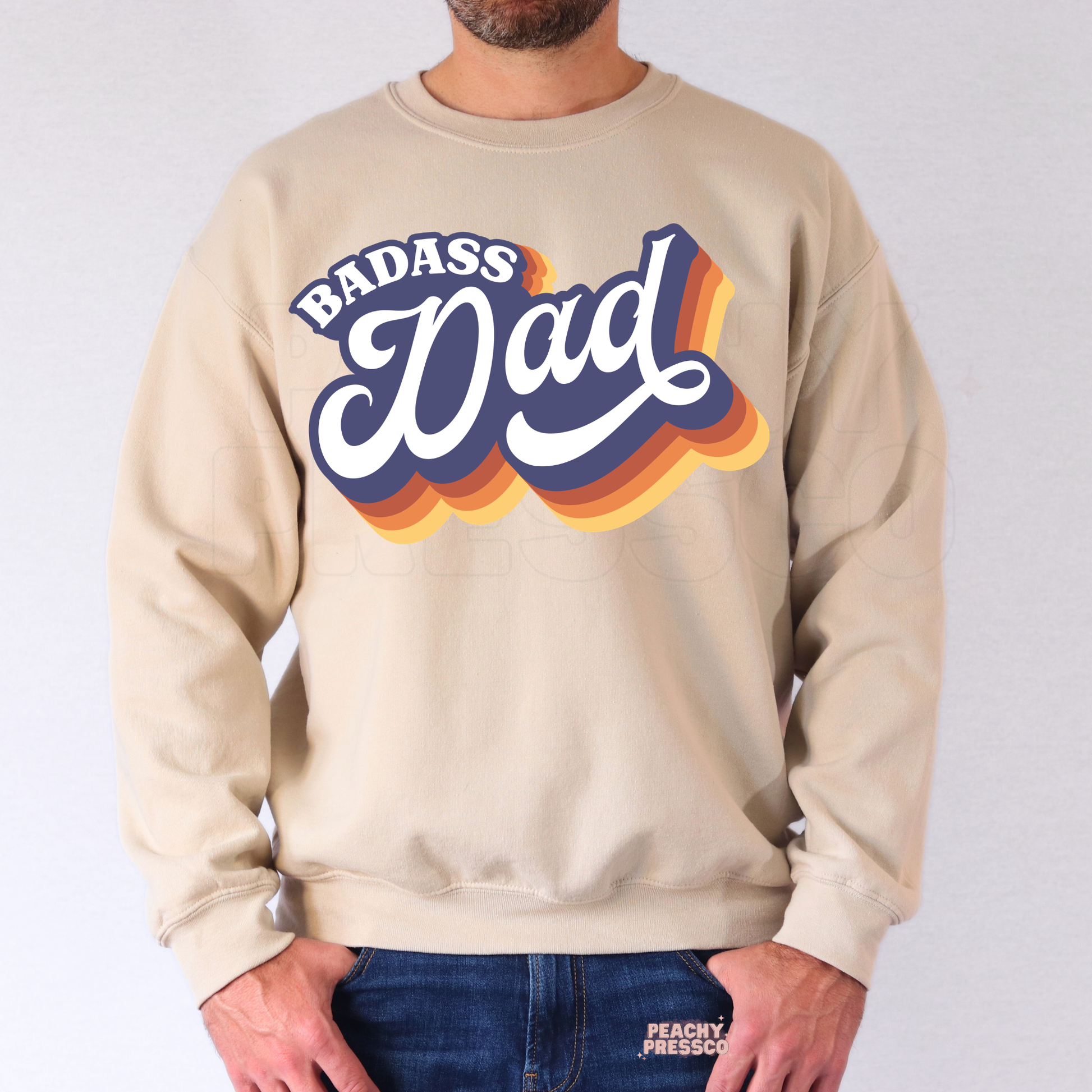 Man modeling beige crewneck sweatshirt featuring colorful retro “Badass Dad” typography graphic for Father’s Day gift.