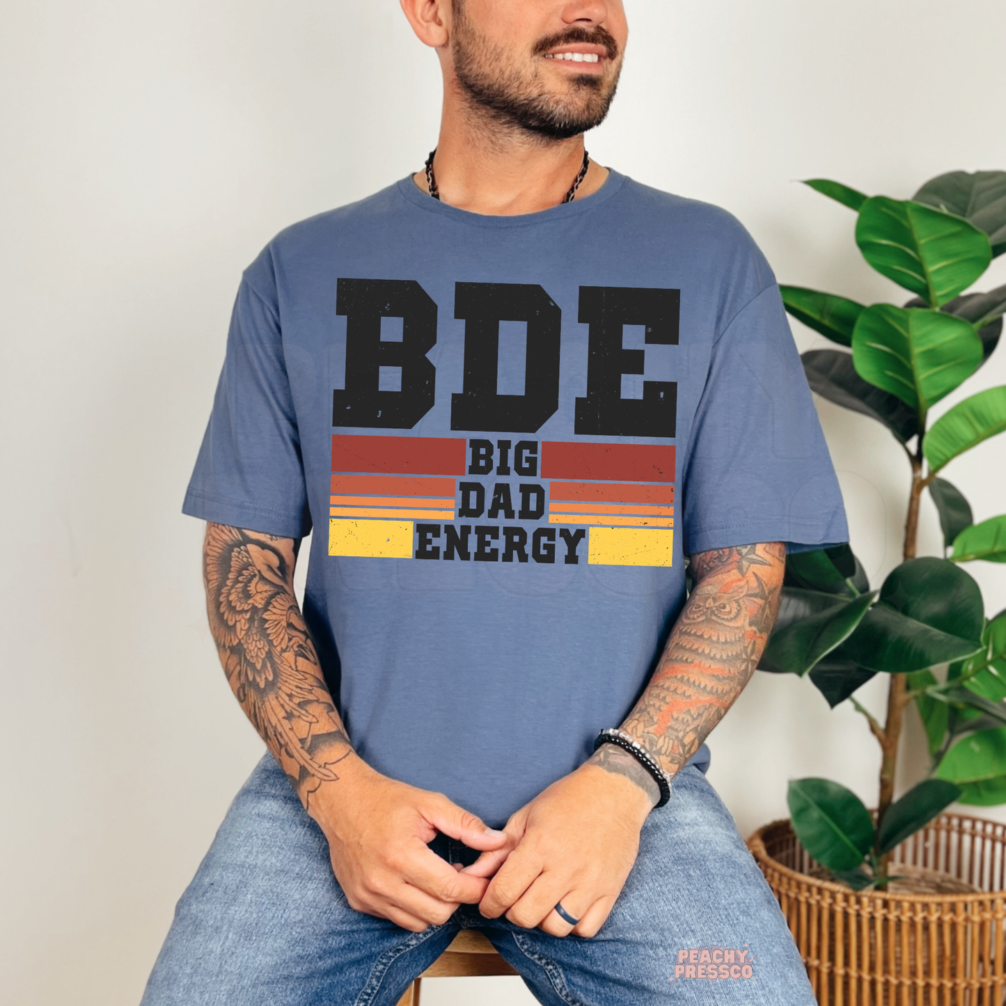 Man wearing blue t-shirt with distressed “BDE Big Dad Energy” graphic in retro sunset colors.