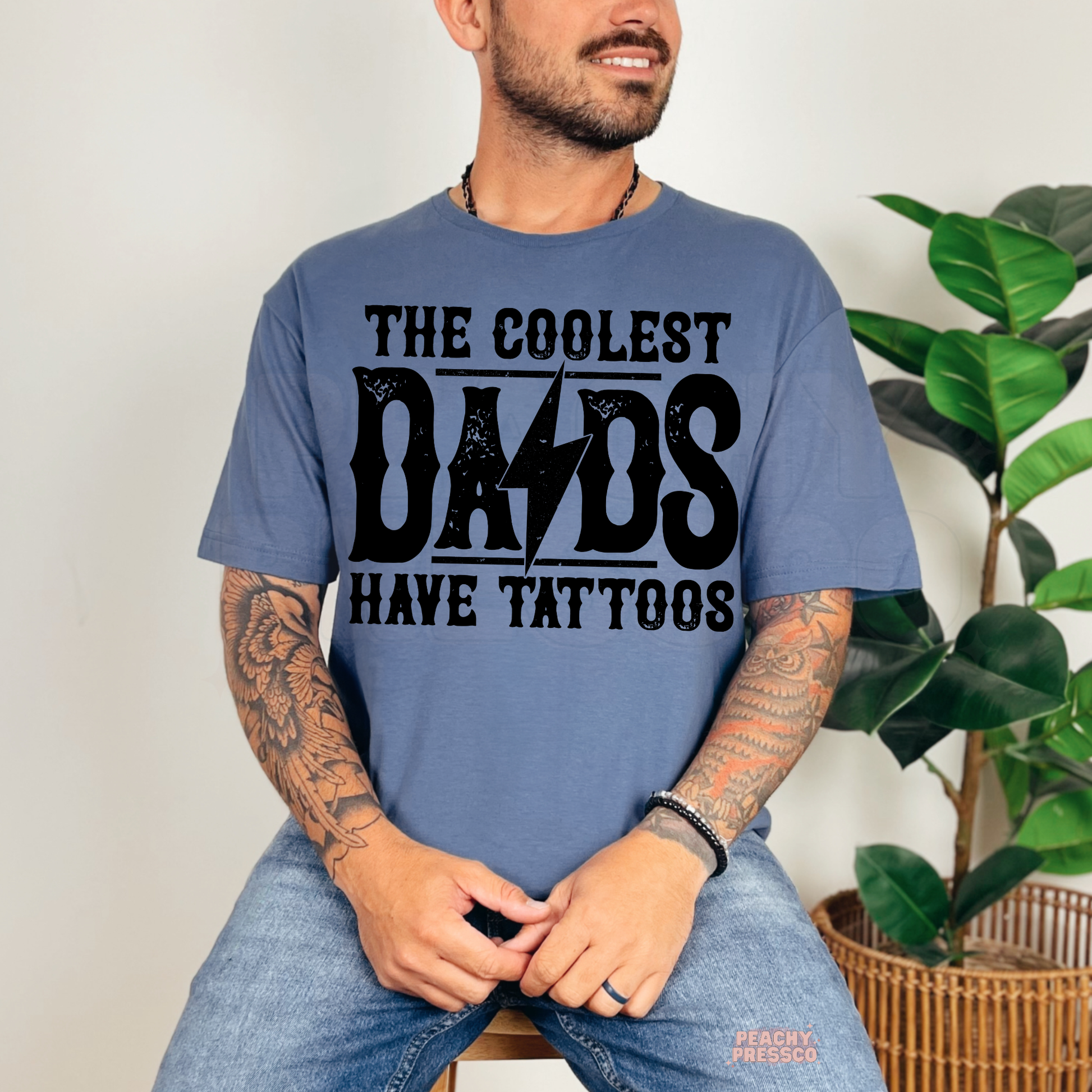 Man modeling blue t-shirt featuring retro “The Coolest Dads Have Tattoos” graphic with lightning bolt design.