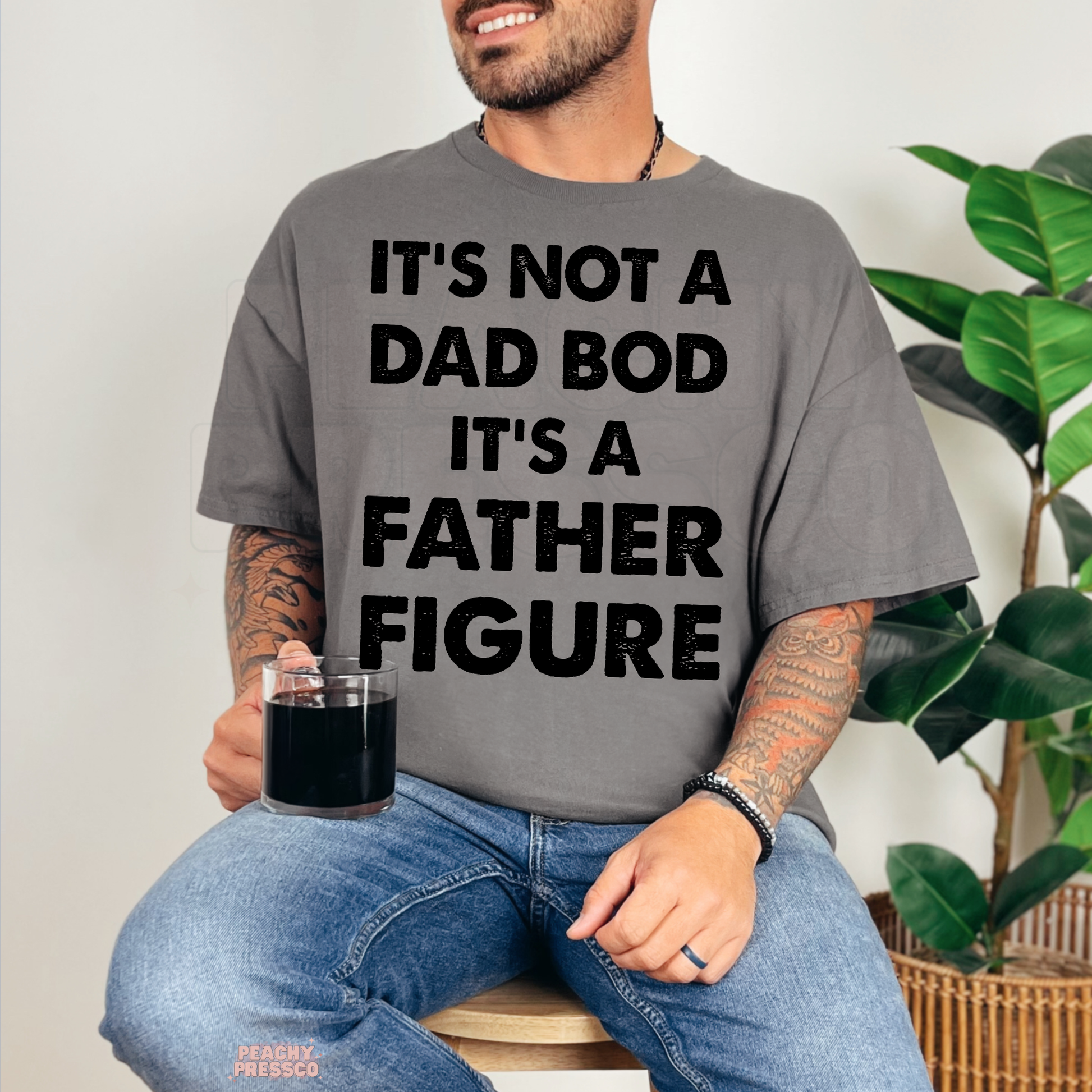 Man sitting with coffee wearing charcoal grey t-shirt featuring humorous “Father Figure” dad graphic print.