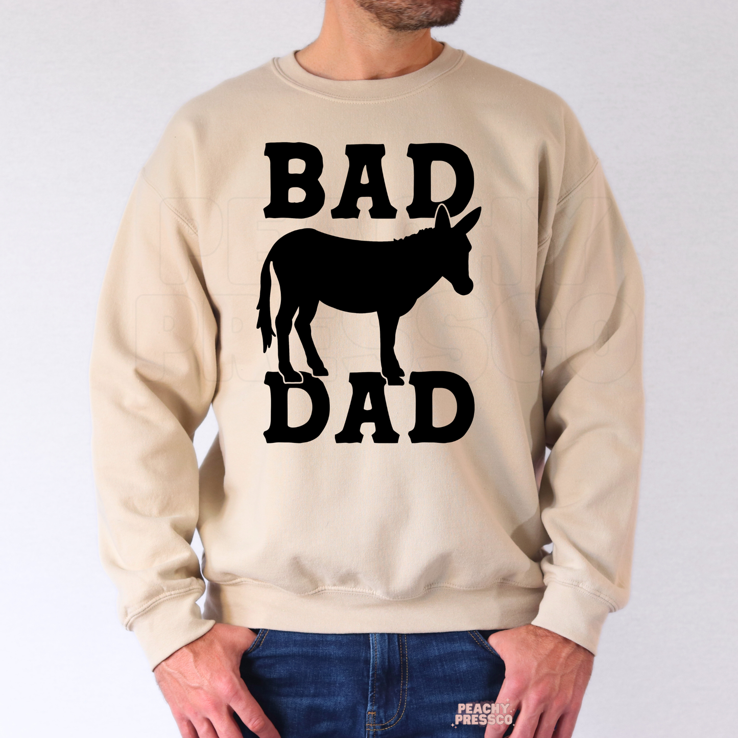 Man wearing beige sweatshirt with black “Bad Dad” western-inspired donkey silhouette graphic.