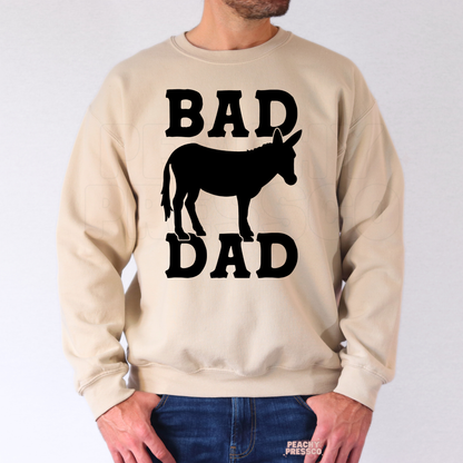 Man wearing beige sweatshirt with black “Bad Dad” western-inspired donkey silhouette graphic.