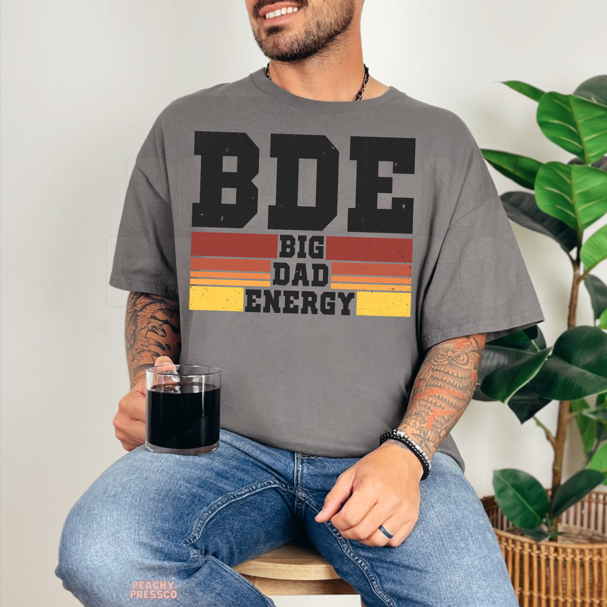 Man sitting with coffee wearing charcoal grey t-shirt featuring retro “BDE Big Dad Energy” dad graphic.