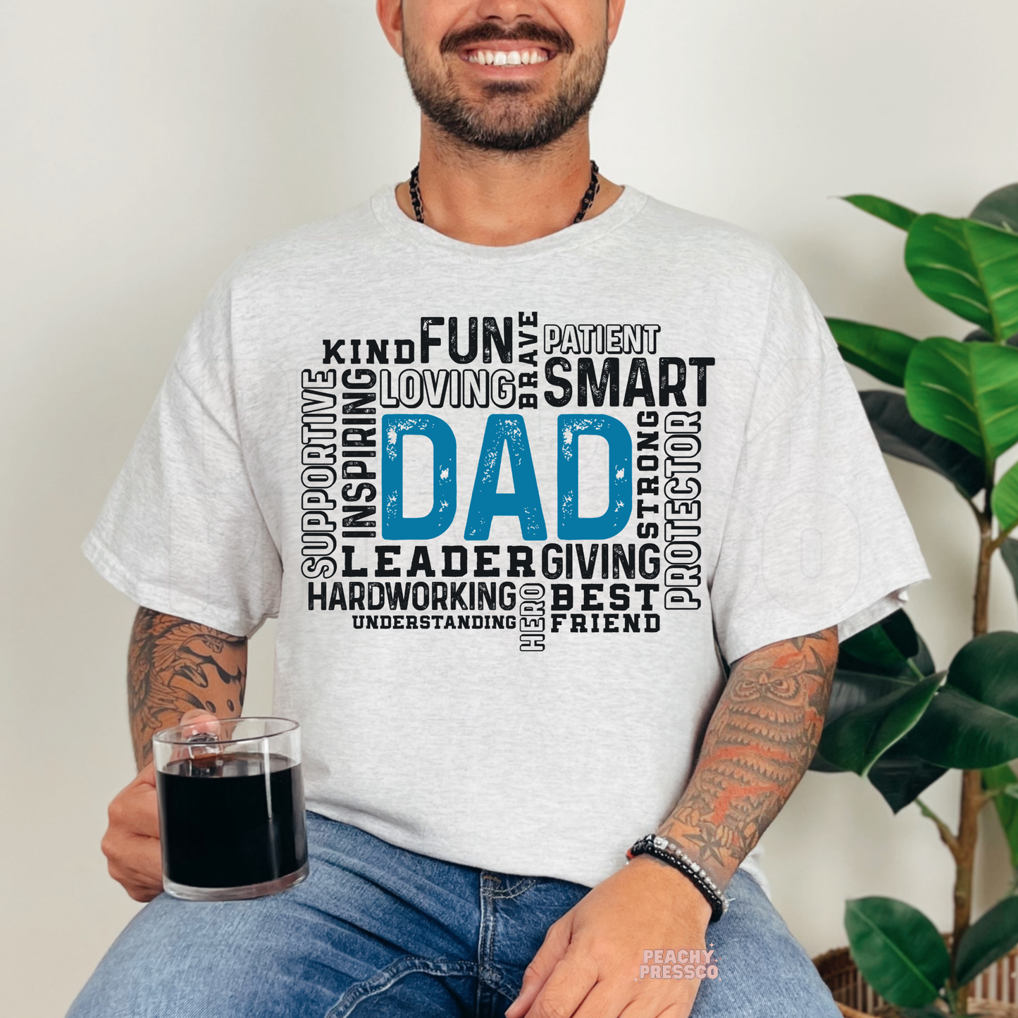 Smiling man wearing ash grey t-shirt with blue and black typography word art design featuring positive traits around the word “Dad.”