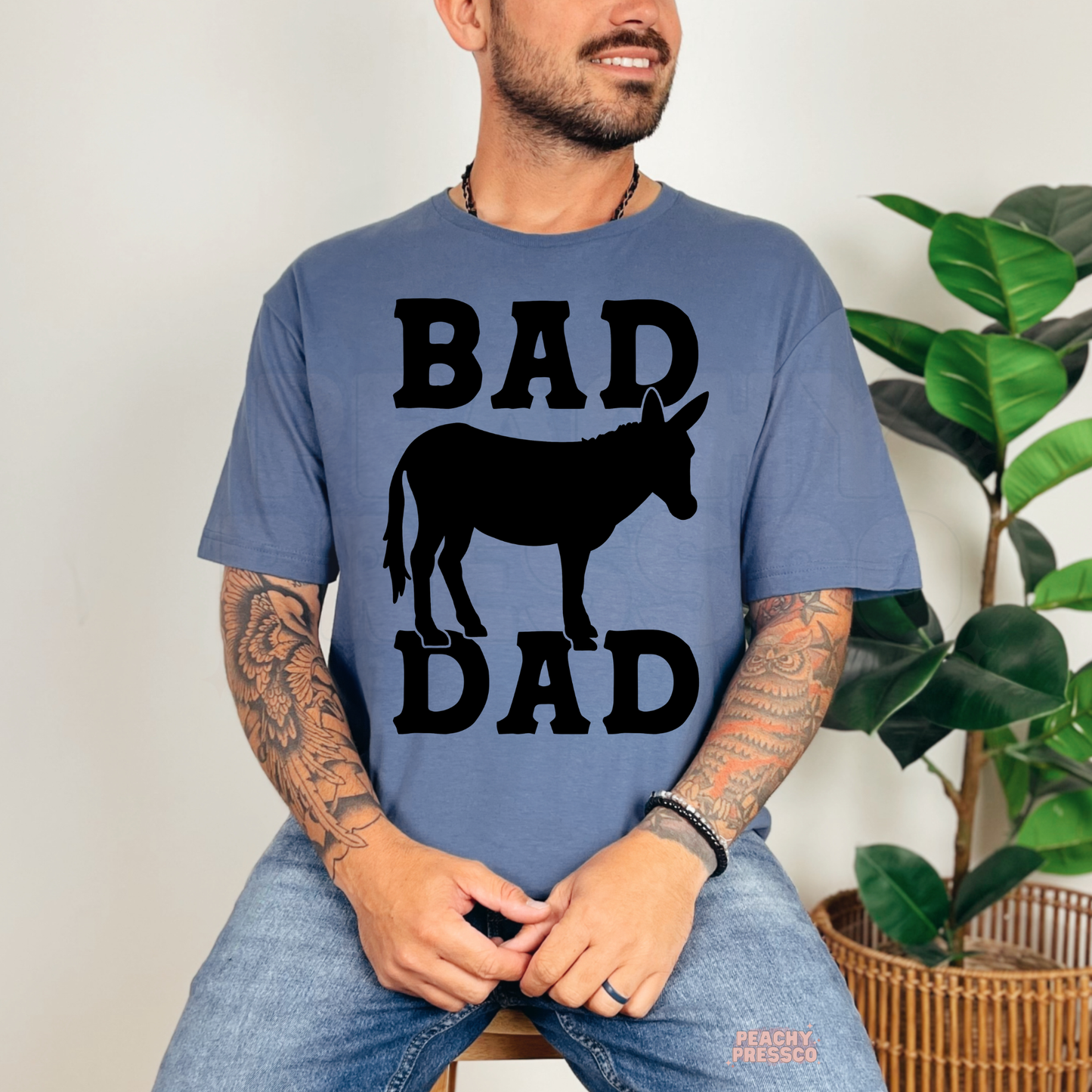 Man wearing faded blue t-shirt with bold black “Bad Dad” donkey silhouette graphic while sitting indoors.