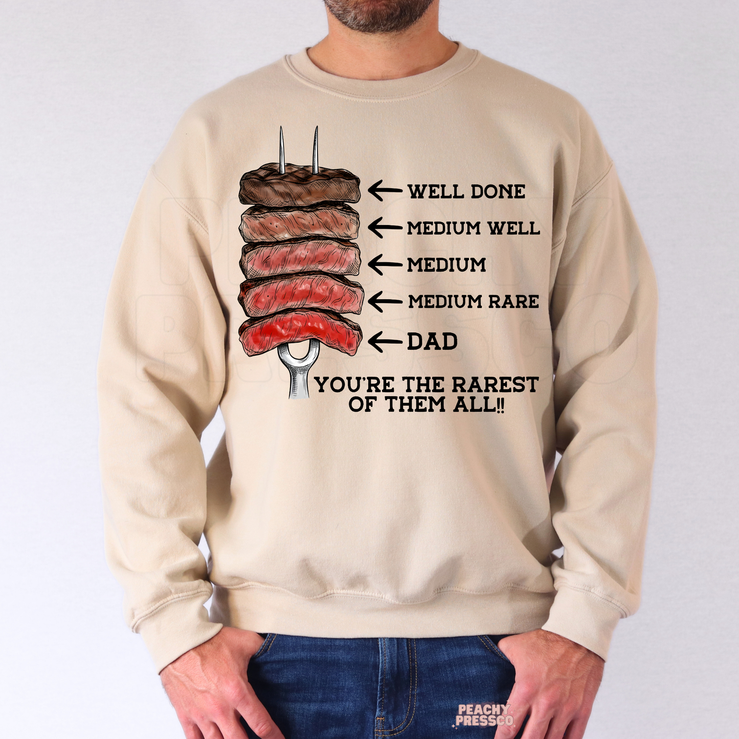 Man wearing beige crewneck sweatshirt with illustrated steak doneness Father’s Day design and “You’re the Rarest of Them All” quote.
