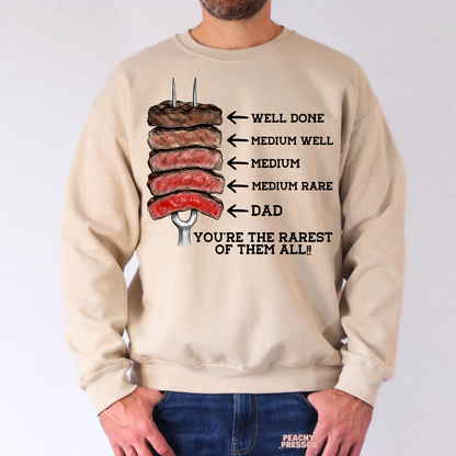 Man wearing beige crewneck sweatshirt with illustrated steak doneness Father’s Day design and “You’re the Rarest of Them All” quote.