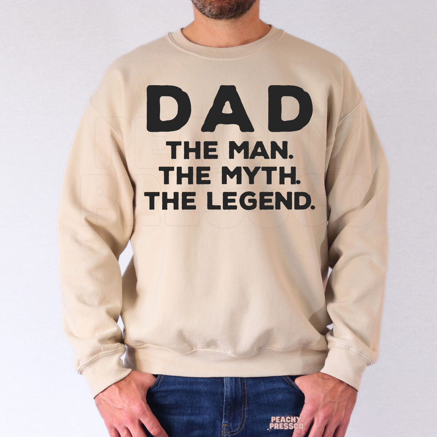 Man modeling sand-colored crewneck sweatshirt with retro-style “Dad The Man. The Myth. The Legend.” graphic print.