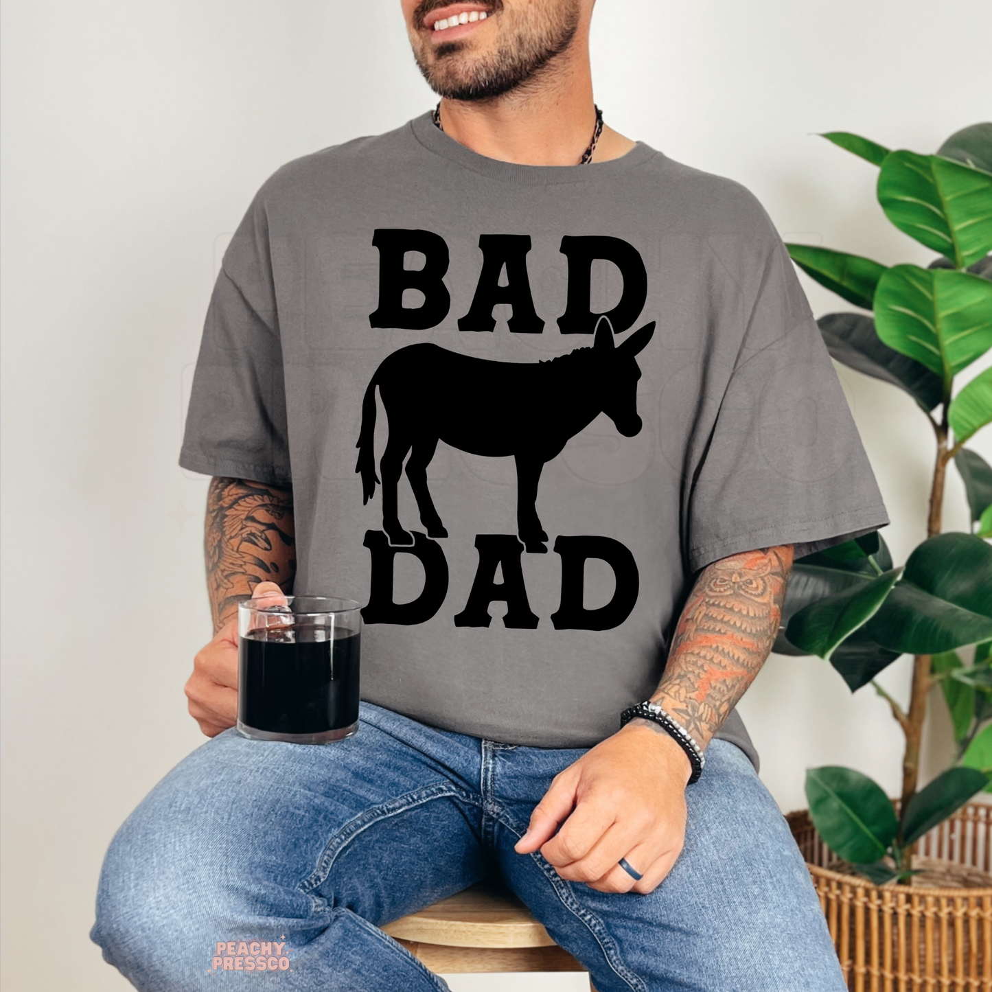 Man wearing charcoal grey t-shirt with bold black “Bad Dad” text and donkey silhouette graphic while sitting with a coffee mug.