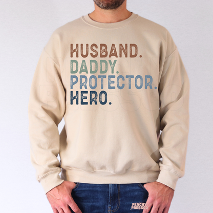 Man modeling sand-colored crewneck sweatshirt featuring distressed “Husband. Daddy. Protector. Hero.” Father’s Day text design.