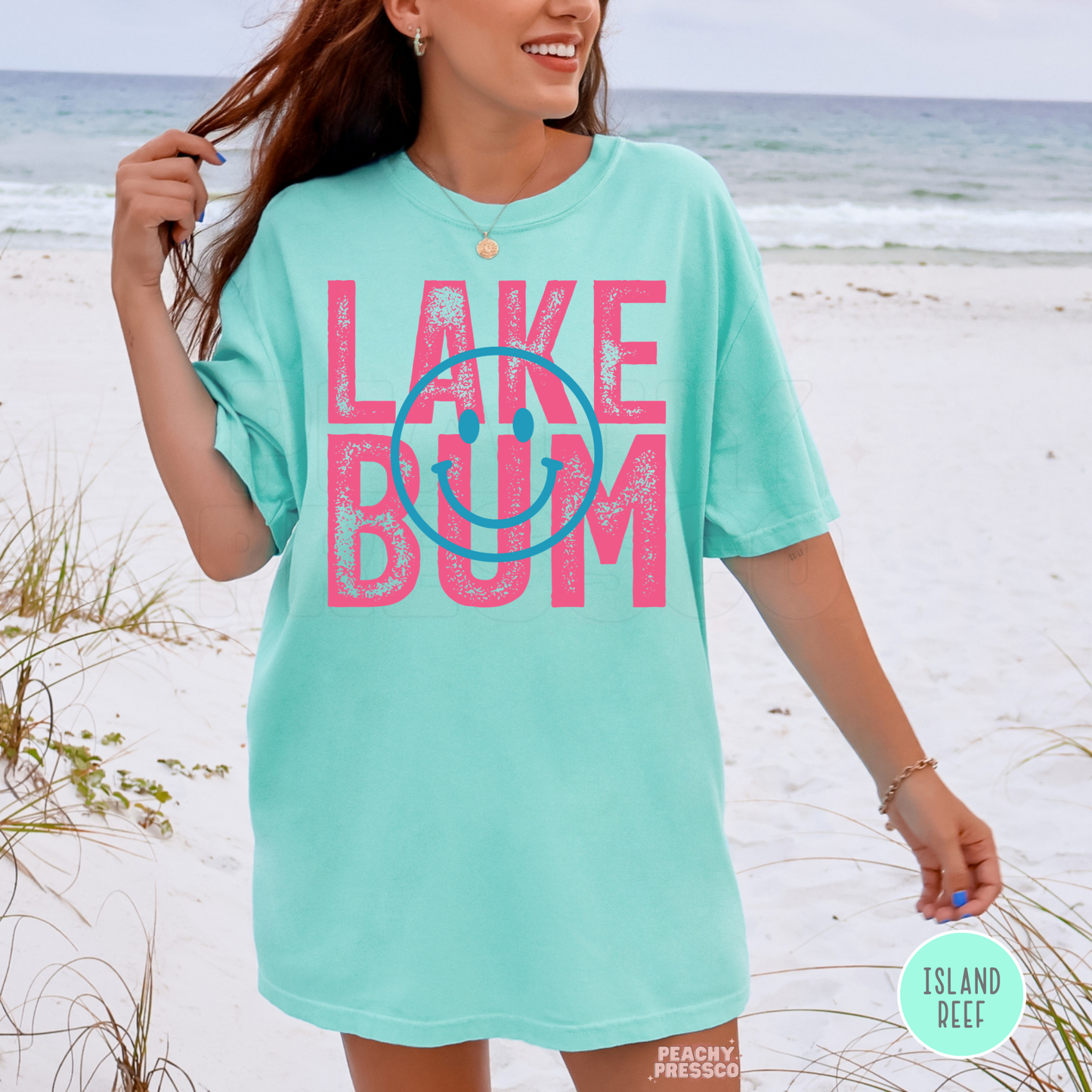 Woman wearing oversized Island Reef Lake Bum graphic t-shirt at the beach with pink retro lettering and blue smiley design.