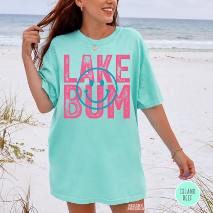 Woman wearing oversized Island Reef Lake Bum graphic t-shirt at the beach with pink retro lettering and blue smiley design.