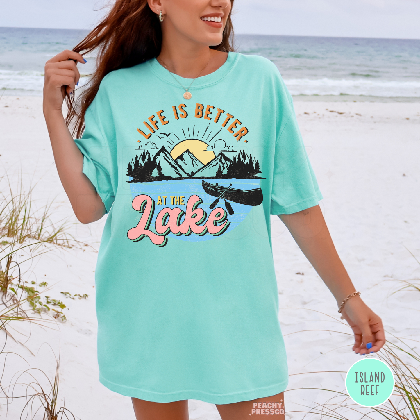 Woman wearing oversized Island Reef graphic t-shirt at the beach featuring “Life Is Better at the Lake” mountain and canoe design.
