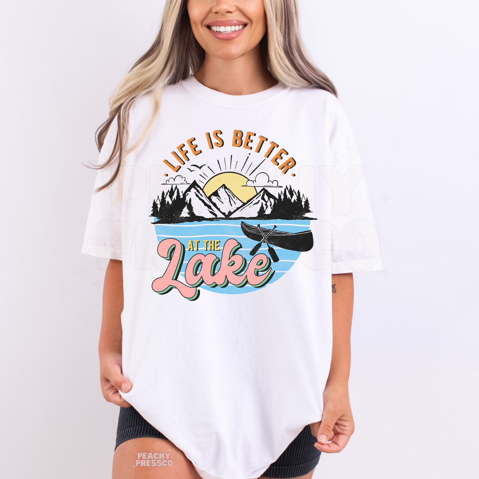 Woman wearing oversized white Life Is Better At The Lake graphic t-shirt with canoe and mountain illustration.