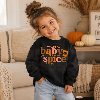 Smiling toddler girl wearing a black "Baby Spice" graphic crewneck sweatshirt with retro fall lettering and pumpkin design in a cozy autumn setting.