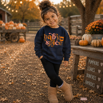 Smiling toddler girl wearing a navy Baby Spice crewneck sweatshirt at a pumpkin patch with pumpkins, autumn leaves, and cozy fall decorations.