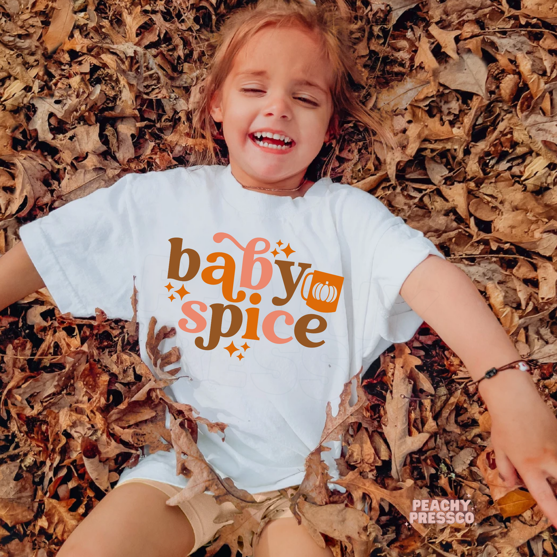 Toddler girl lying in colorful autumn leaves wearing a white "Baby Spice" graphic t-shirt with retro fall lettering and pumpkin accent.