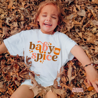 Toddler girl lying in colorful autumn leaves wearing a white "Baby Spice" graphic t-shirt with retro fall lettering and pumpkin accent.