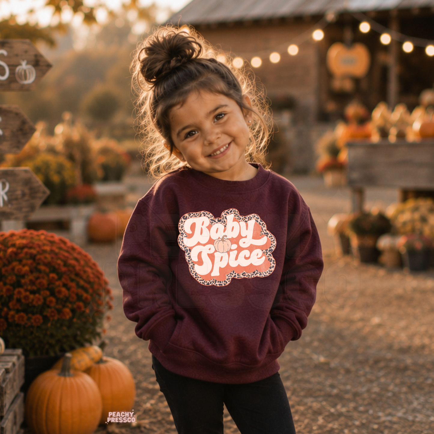 Toddler girl smiling in a maroon "Baby Spice" sweatshirt with a retro leopard print design during a cozy autumn pumpkin patch visit.