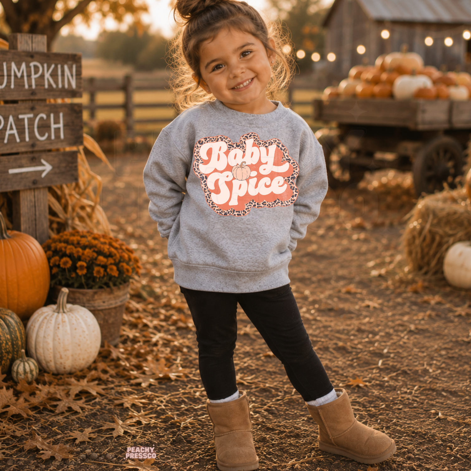 Happy toddler girl wearing a sport grey "Baby Spice" sweatshirt with a retro pumpkin-inspired graphic at a rustic pumpkin patch.