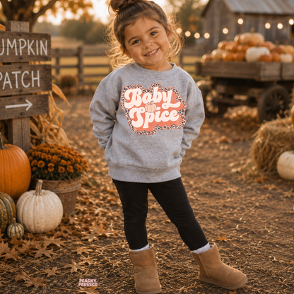 Happy toddler girl wearing a sport grey "Baby Spice" sweatshirt with a retro pumpkin-inspired graphic at a rustic pumpkin patch.