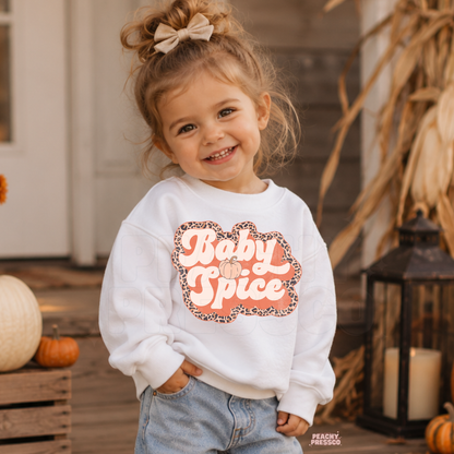 Smiling toddler girl wearing a white "Baby Spice" sweatshirt with a retro leopard print design and pumpkin graphic on a cozy fall porch.