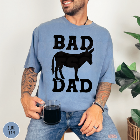 Man wearing a blue jean garment-dyed t-shirt featuring a funny "Bad Ass Dad" donkey graphic with bold western lettering.