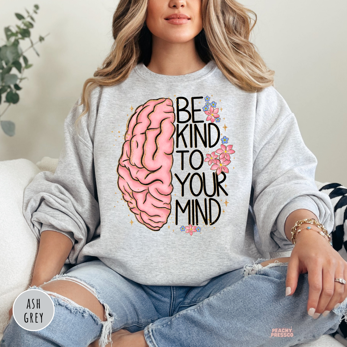 Ash grey sweatshirt featuring a pink brain illustration with floral accents and the inspirational quote "Be Kind to Your Mind" in a modern hand-lettered design.