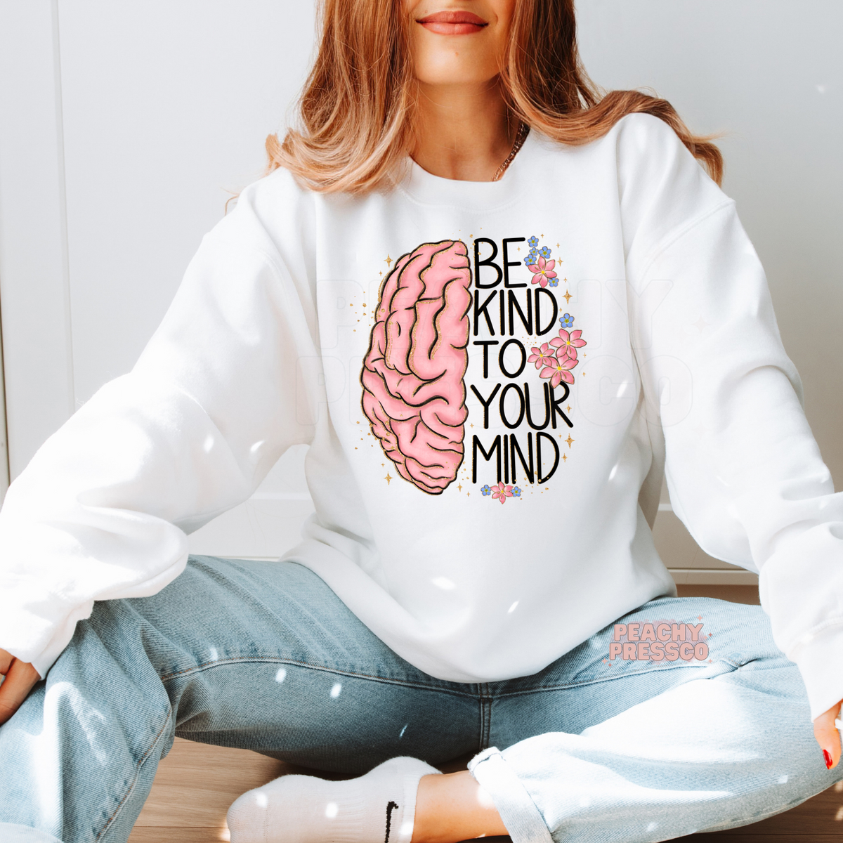 White sweatshirt displaying a pink brain graphic with floral accents and the uplifting message "Be Kind to Your Mind" for mental wellness and positivity.