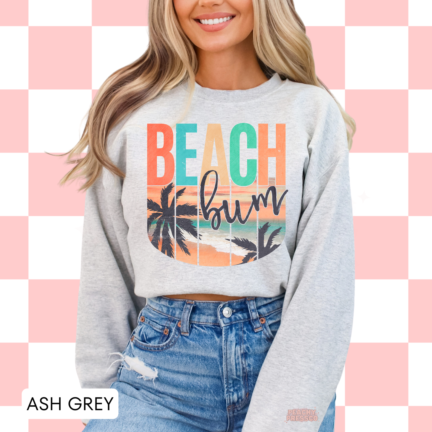 Woman wearing an ash grey crewneck sweatshirt featuring a retro "Beach Bum" tropical sunset graphic with palm trees, beach shoreline, and vintage coastal colors.