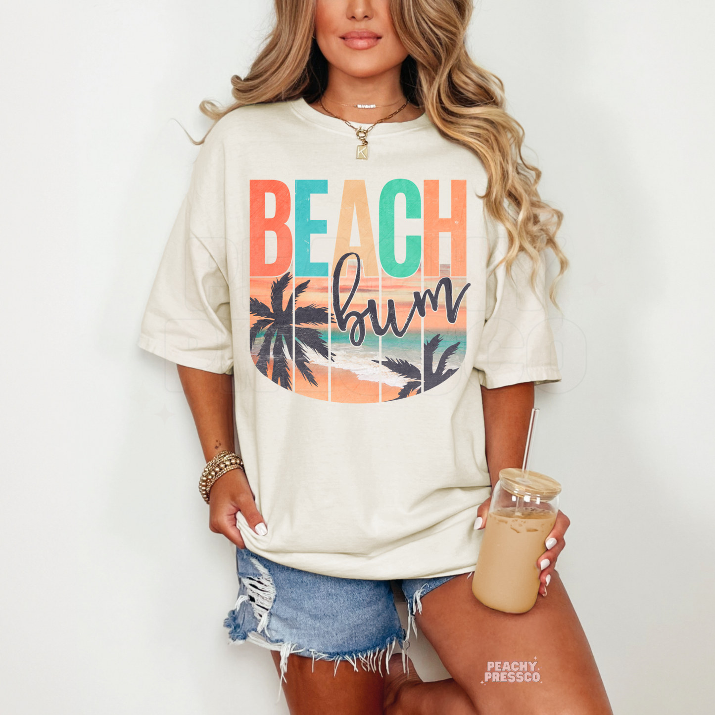 Woman wearing an ivory t-shirt featuring a retro "Beach Bum" beach graphic with colorful sunset, palm tree silhouettes, ocean shoreline, and vintage coastal style.
