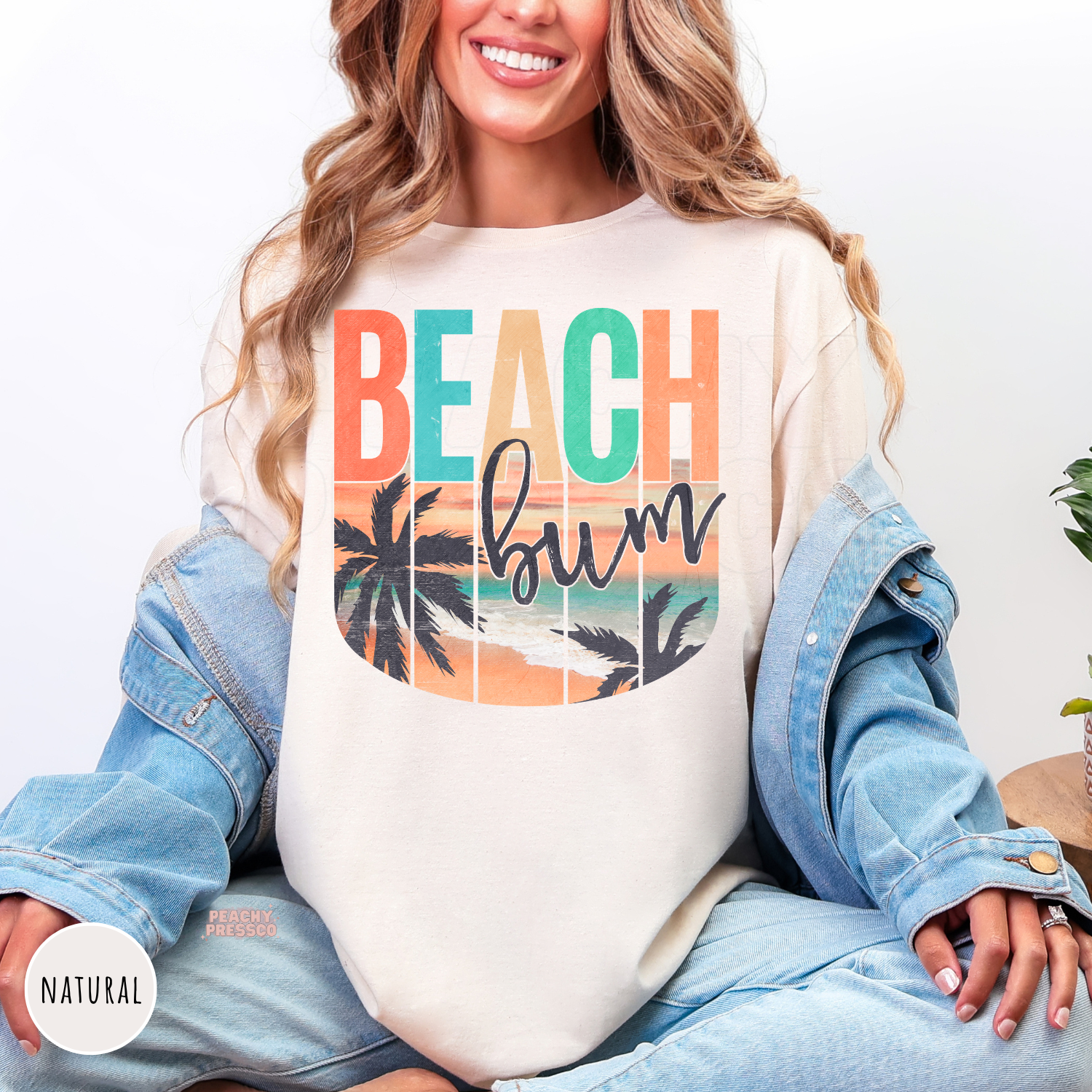 Woman wearing a natural colored tshirt featuring a retro "Beach Bum" beach design with palm trees, ocean sunset, and vintage-inspired tropical artwork.