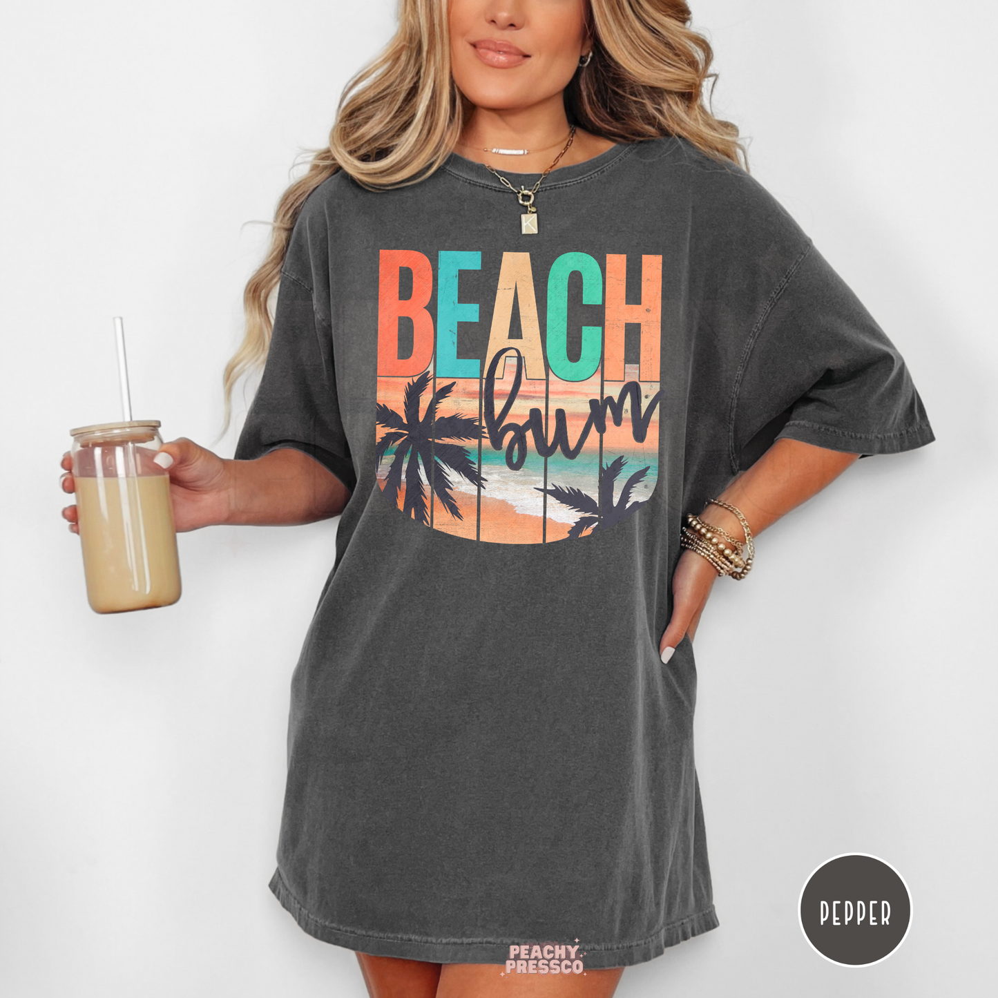 Woman wearing a pepper Comfort Colors t-shirt featuring a retro "Beach Bum" sunset design with palm trees, ocean waves, and vintage tropical beach colors.