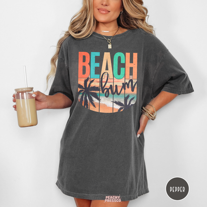 Woman wearing a pepper Comfort Colors t-shirt featuring a retro "Beach Bum" sunset design with palm trees, ocean waves, and vintage tropical beach colors.