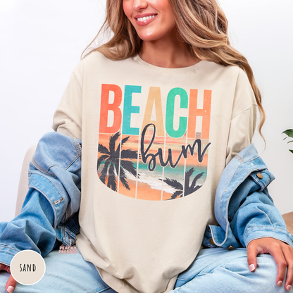 Woman wearing a sand colored crewneck sweatshirt featuring a retro "Beach Bum" beach sunset graphic with palm trees, ocean shoreline, and colorful vintage tropical design.
