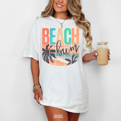 Woman wearing a white t-shirt featuring a retro "Beach Bum" beach sunset design with palm trees, ocean waves, and colorful vintage summer artwork.