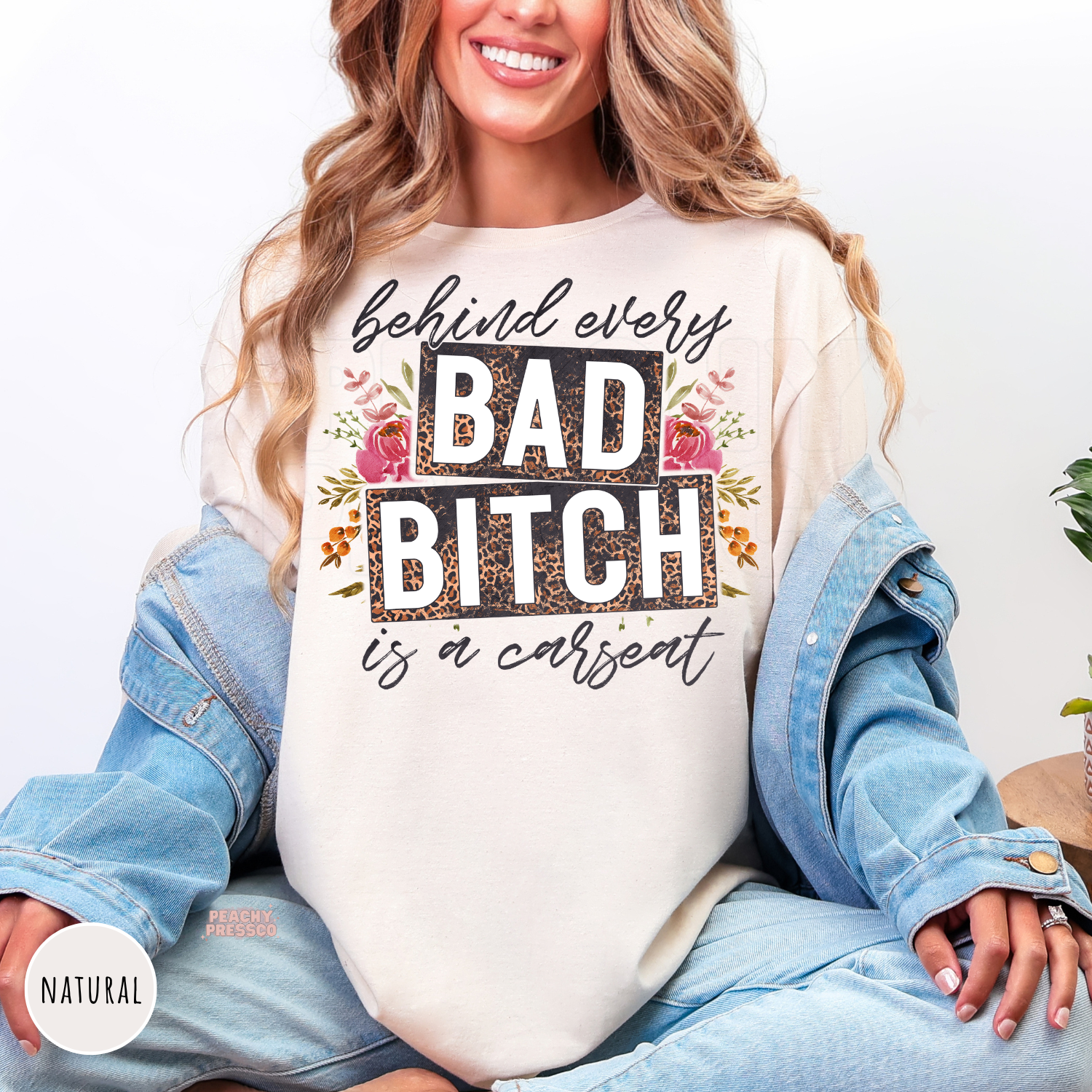Woman wearing a natural shirt with behind every bad bitch is a carseat graphic featuring leopard print blocks and floral accents.