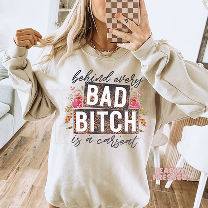 Mirror selfie of woman wearing a sand sweatshirt with behind every bad bitch is a carseat graphic in leopard print with floral accents.