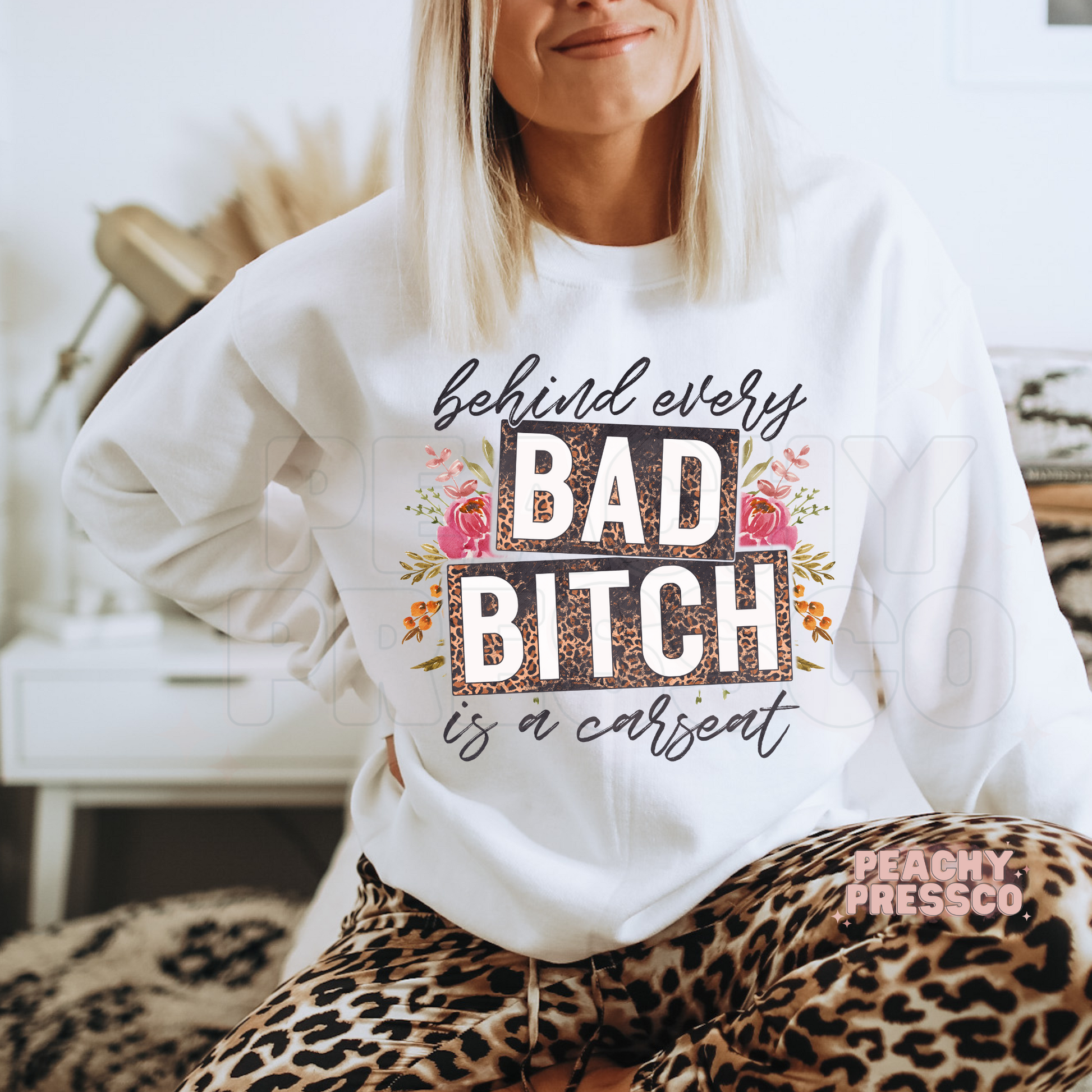 Woman relaxing in a white sweatshirt with behind every bad bitch is a carseat funny motherhood graphic and floral leopard print design.