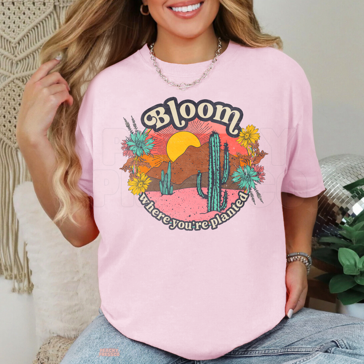 Light pink Bloom Where You're Planted graphic shirt featuring a retro desert sunset, green cacti, colorful wildflowers, and inspirational botanical quote.