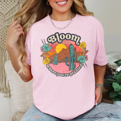 Light pink Bloom Where You're Planted graphic shirt featuring a retro desert sunset, green cacti, colorful wildflowers, and inspirational botanical quote.