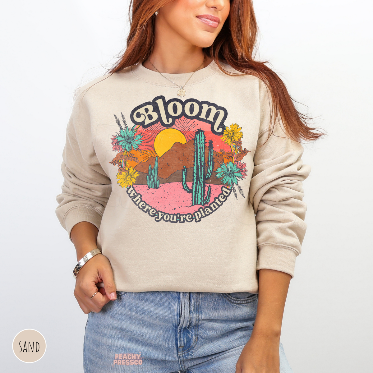 Sand Bloom Where You're Planted sweatshirt showcasing a colorful retro desert scene with tall green cacti, yellow wildflowers, mountains, and sunset.