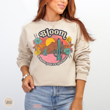 Sand Bloom Where You're Planted sweatshirt showcasing a colorful retro desert scene with tall green cacti, yellow wildflowers, mountains, and sunset.