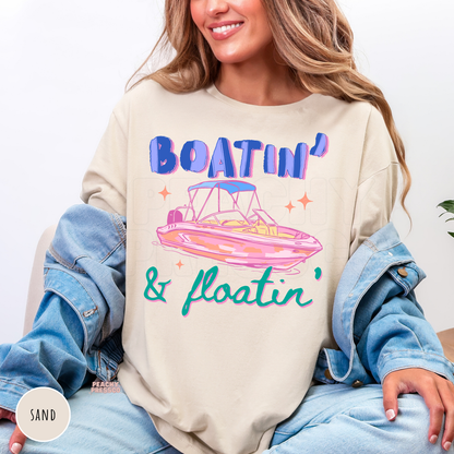 Sand crewneck sweatshirt featuring a colorful retro "Boatin' & Floatin'" graphic with a pink pontoon boat, bold blue lettering, and teal script for lake lovers and boating adventures.