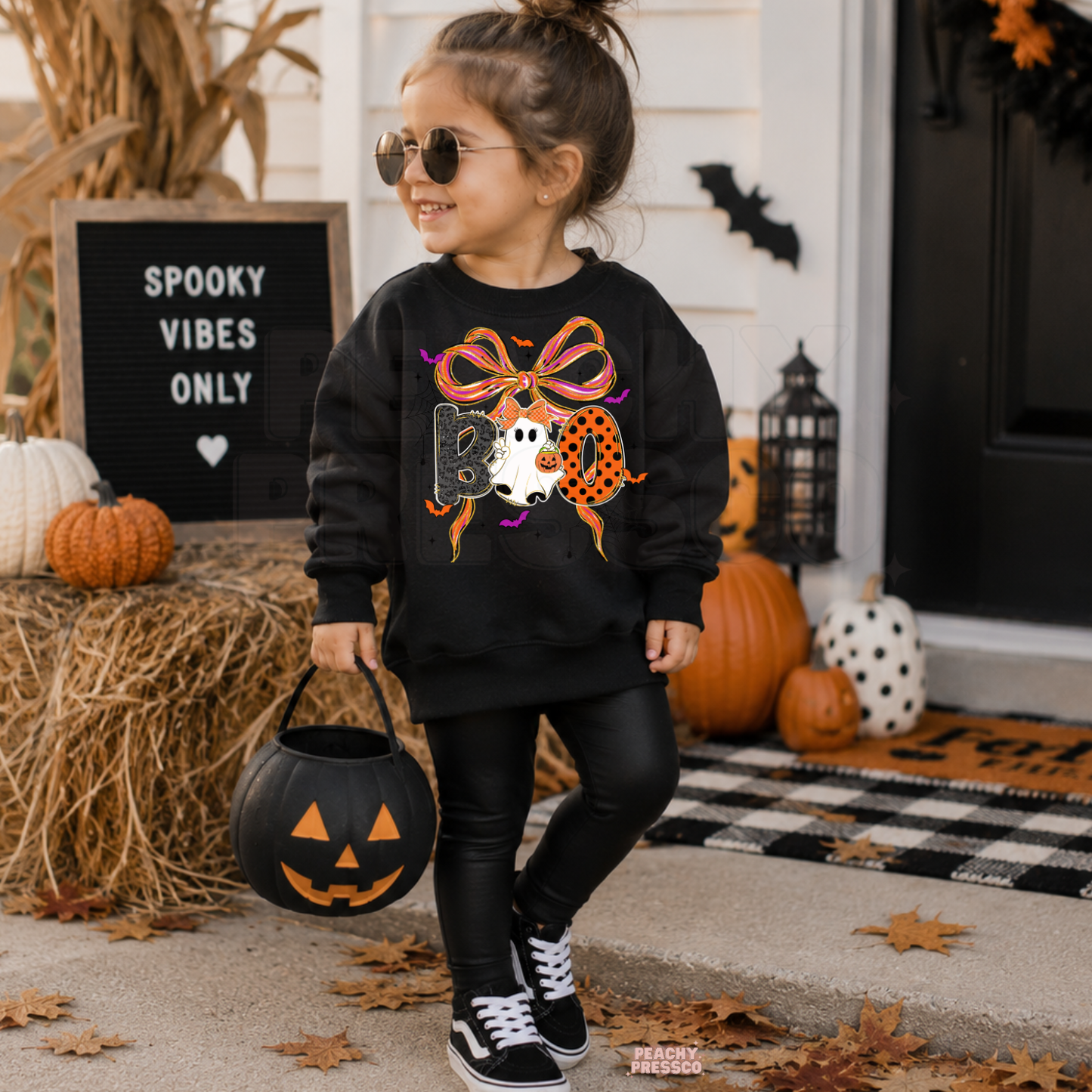 Girl wearing a black Halloween sweatshirt featuring a colorful BOO ghost and bow graphic while holding a pumpkin trick-or-treat bucket.