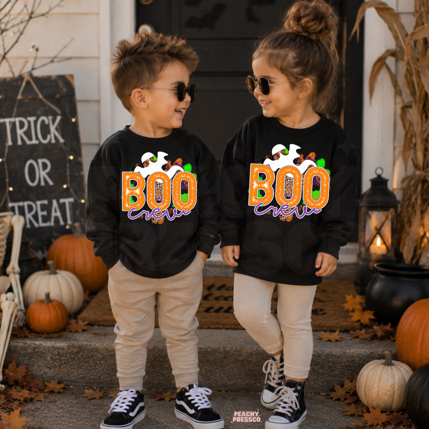 Boy and girl wearing matching black Boo Crew Halloween sweatshirts featuring a colorful ghost design with pumpkins and spooky porch decor.
