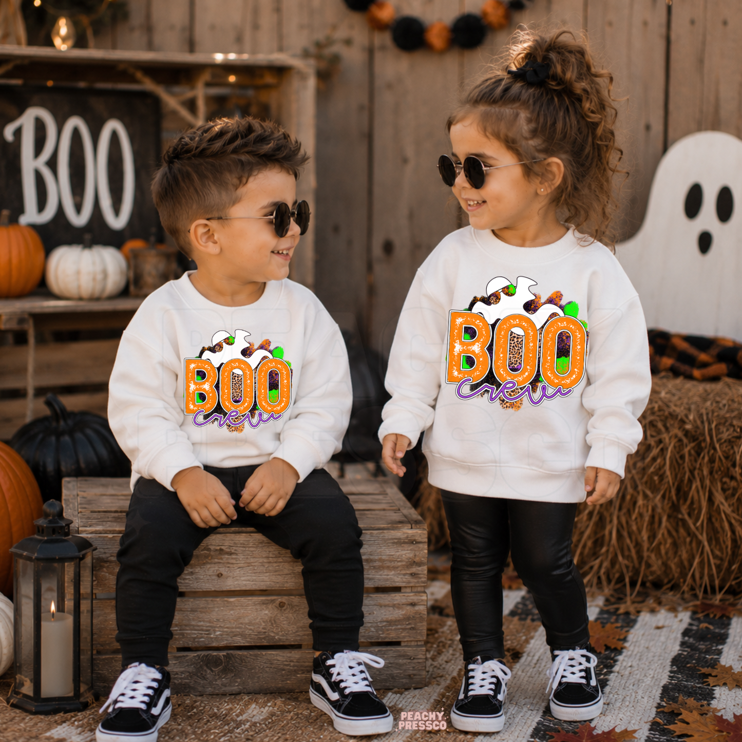 Toddler boy and girl wearing matching white Boo Crew Halloween sweatshirts with a colorful ghost graphic in a festive Halloween setting.