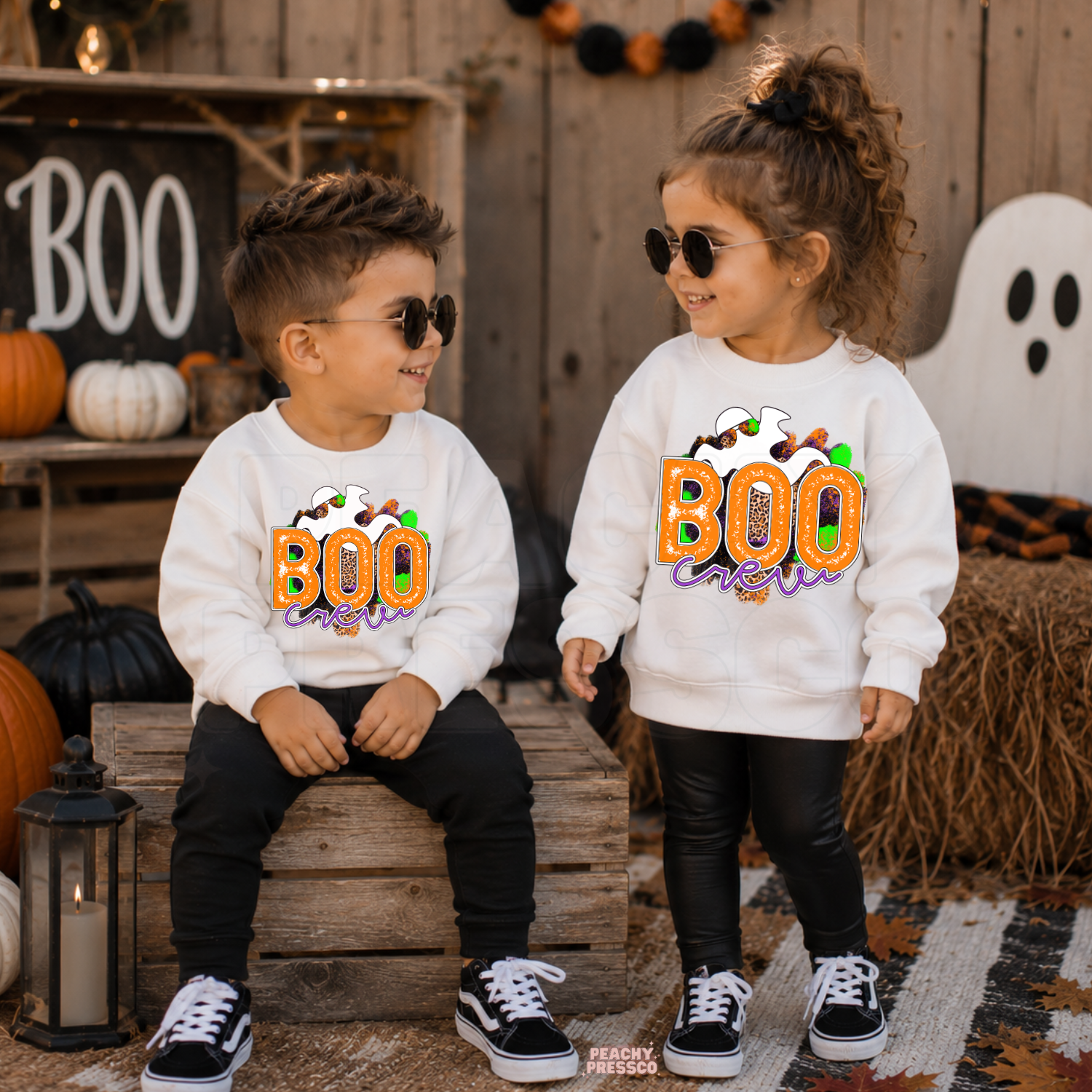 Toddler boy and girl wearing matching white Boo Crew Halloween sweatshirts with a colorful ghost graphic in a festive Halloween setting.