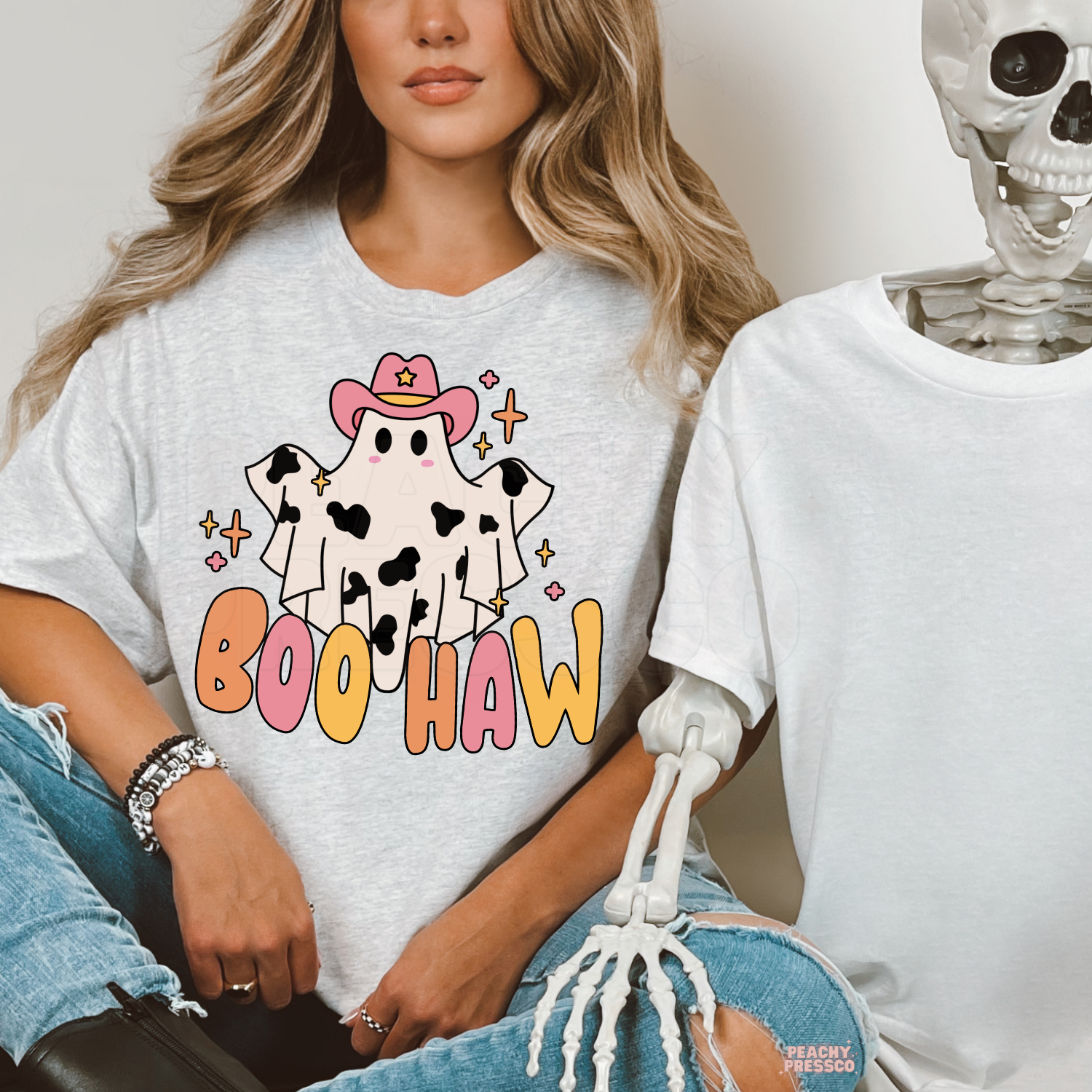 Woman wearing an ash grey Boo Haw Halloween graphic t-shirt with a cute cowgirl ghost beside a Halloween skeleton.